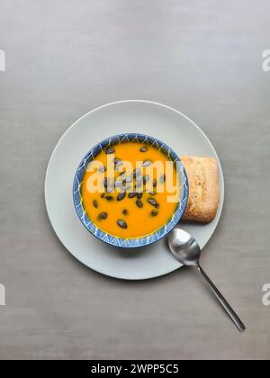 Zuppa di zucca fresca fatta in casa guarnita con semi di zucca e olio di lino come condimento in una ciotola per zuppa su un piatto bianco con cucchiaio e panino Foto Stock