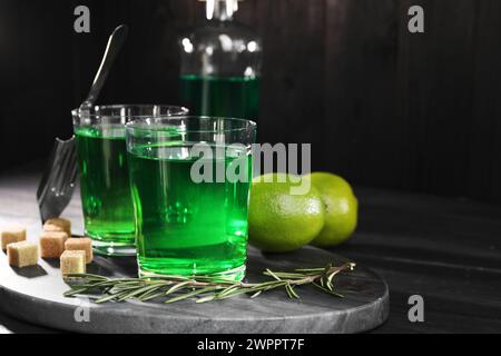 Absinthe in bicchieri, rosmarino, zucchero di canna e lime su un tavolo in legno nero, spazio per il testo. Bevanda alcolica Foto Stock