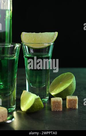 Absinthe in bicchieri da shot, lime e zucchero di canna a cubetti su tavolo grigio su sfondo nero. Bevanda alcolica Foto Stock