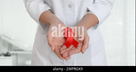 Consapevolezza del cancro. Medico che regge il nastro rosso su sfondo sfocato, primo piano. Design banner Foto Stock