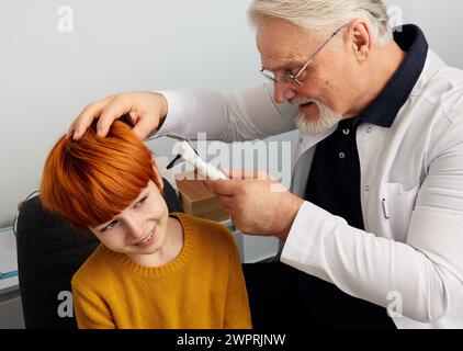 Positivo bambino di sesso maschile con i capelli rossi durante l'esame dell'orecchio presso la clinica dell'udito. Medico ENT che controlla l'orecchio maschile con l'otoscopio Foto Stock