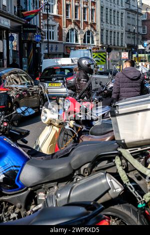 Soho, Londra, Regno Unito, 8 marzo 2024, gli scooter a motore parcheggiato e la linea di traffico su strada trafficata hanno ritardato due persone che parlavano uno indossando un casco antiurto Foto Stock
