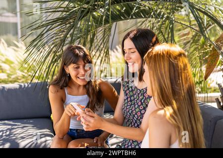 Vista laterale di tre bellissime donne che guardano qualcosa sullo smartphone mentre sono sedute su un divano Foto Stock