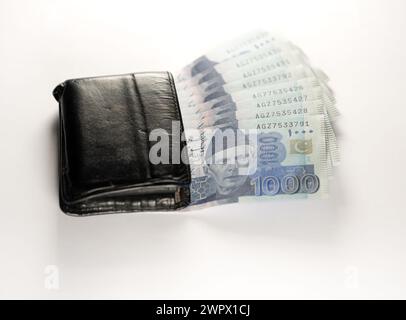 Mille banconote da 1000 rupie allineate a un portafoglio nero in pelle. Banconote ufficiali della Banca statale pakistana 2024 isolate su sfondo bianco. Foto Stock