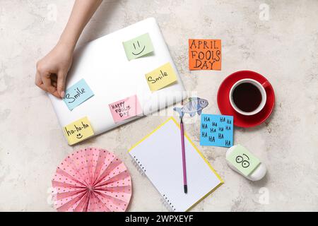 Mano femminile che attacca la carta con UN SORRISO di testo vicino al computer portatile e tazza di caffè su sfondo grunge. April Fools Day Pank Foto Stock