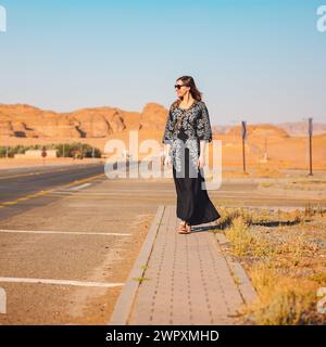 Giovane donna con lungo abito nero che cammina sul marciapiede vicino alla strada - paesaggio roccioso del deserto, tipico di Alula, sullo sfondo Foto Stock