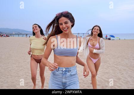 Tre donne in costume da bagno corrono lungo la spiaggia sorridendo alla macchina fotografica, divertendosi durante le vacanze estive. Foto Stock