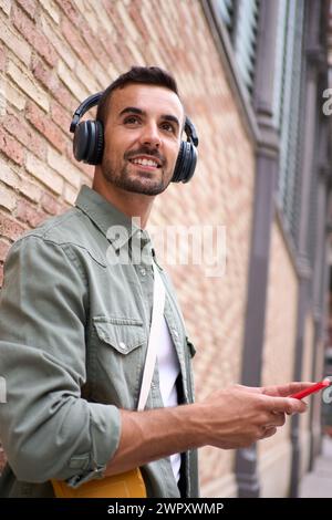 Giovane e affascinante uomo sorridente con le cuffie wireless che reggono il cellulare appoggiato contro il muro di mattoni. Foto Stock