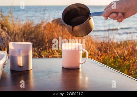 l'uomo versa il caffè appena estratto da un mestolo in alluminio in una tazza su un tavolo turistico, in riva al mare. Viaggi e turismo. Foto Stock