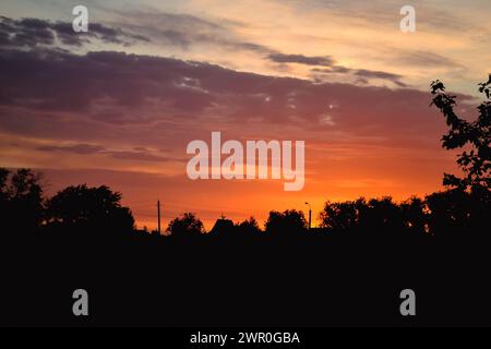 Il bagliore mattutino illumina le nuvole su un bellissimo cielo colorato sopra le sagome degli alberi, una stufa e un tetto triangolare di una casa all'alba Foto Stock