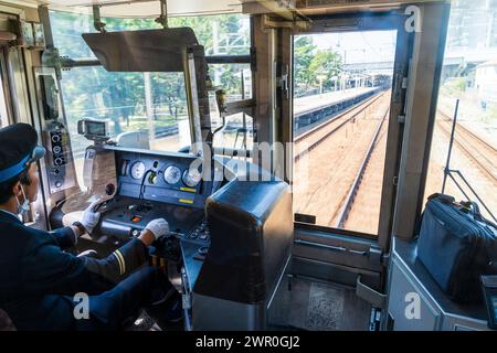 Vista frontale attraverso la cabina del conducente di un treno pendolare con l'autista che guida mentre accelera attraverso una stazione cittadina sulla linea JR Kobe in Giappone. Foto Stock