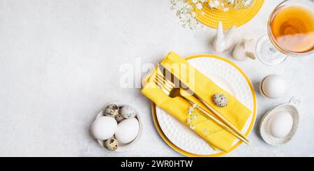 Il tavolo del ristorante bianco e giallo serve vista dall'alto. Uova, piatti bianchi e gialli, posate dorate, bicchiere di vino rosa su bianco. Tavola per Pasqua Foto Stock