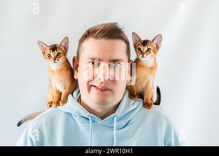 Uomo divertente sorridente con felpa blu con cappuccio e gemelli gatti abissini, piccoli gattini appena nati seduti sulle spalle. Sfondo bianco. Animali domestici felici, soffici domestici Foto Stock