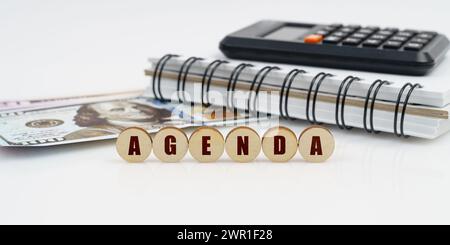 Business e finanza. Su una superficie alta si trovano un blocco note, una calcolatrice, dollari e cerchi di legno con l'iscrizione - AGENDA Foto Stock