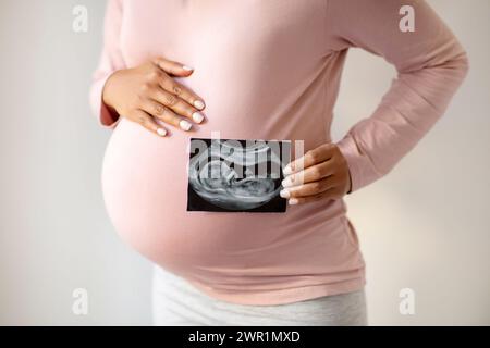 Donna incinta che mostra amorevolmente la sua foto ecografica e abbraccia dolcemente la pancia Foto Stock