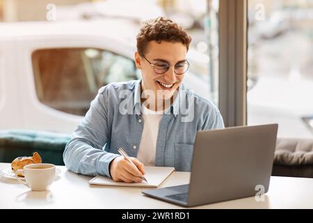 Allegro giovane uomo dai capelli ricci che indossa occhiali e scrive su un notebook con un caffè Foto Stock