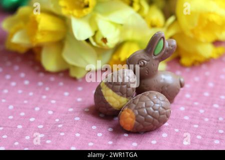 Coniglietti e narcisi al cioccolato pasquale su tovaglia rosa. Cartolina di Pasqua luminosa e colorata. Foto Stock