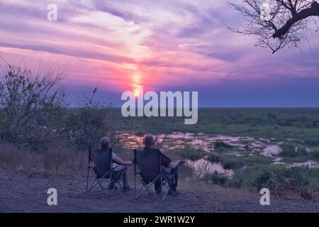Coppia seduta su sedie vicino a un fiume in Africa a guardare il sole tramontare sulla pianura africana. Foto Stock