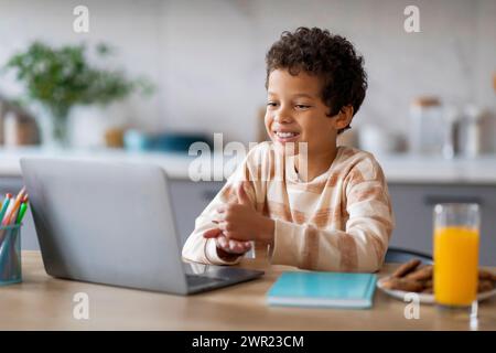 Felice ragazzo nero che interagisce con il computer portatile seduto al tavolo in cucina Foto Stock