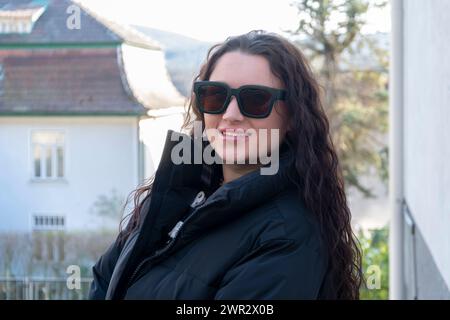 Una donna felice con lunghi capelli ricci sta all'esterno indossando occhiali da sole e una giacca nera sullo sfondo della campagna. Foto Stock