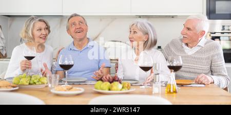 Due coppie sposate mature sono sedute a tavola, gustando cibo e vino, chiacchierando. Foto Stock