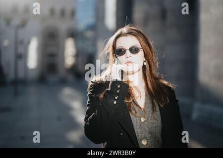 Una giovane donna d'affari con occhiali da sole alla moda si impegna in una conversazione telefonica seria, illuminata dalla luce ambientale della città. Foto Stock