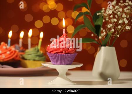 Mettiti in piedi con un cupcake di compleanno sul tavolo bianco contro luci sfocate Foto Stock