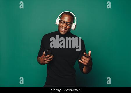 Il giovane con occhiali e abbigliamento casual balla con gioia su sfondo verde. Ha in mano uno smartphone e indossa un auricolare elegante per ascoltare Foto Stock