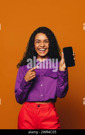 Una donna allegra con capelli ricci si staglia su un vivace sfondo arancione, tenendo in mano un dispositivo mobile. Guarda la telecamera, sorridendo, mentre lo consiglia Foto Stock