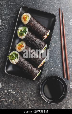 Un piatto nero con pezzi di sushi con bacchette e ciotola di salsa di soia. Il sushi mostra una varietà di colori, texture e ripieni, in vetrina Foto Stock