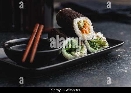 Un piatto nero con pezzi di sushi con bacchette e ciotola di salsa di soia. Il sushi mostra una varietà di colori, texture e ripieni, in vetrina Foto Stock