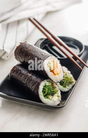Un piatto pieno di una varietà di sushi roll con un set di bacchette di legno e una ciotola di salsa di soia, collocata su un semplice tavolo da cucina. Foto Stock