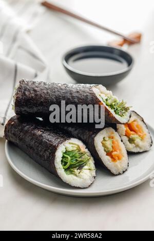 Un piatto pieno di una varietà di sushi roll con un set di bacchette di legno e una ciotola di salsa di soia, collocata su un semplice tavolo da cucina. Foto Stock
