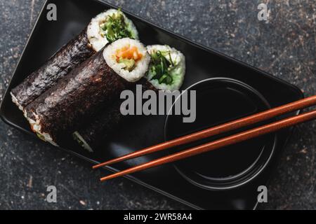 Un piatto nero con pezzi di sushi con bacchette e ciotola di salsa di soia. Il sushi mostra una varietà di colori, texture e ripieni, in vetrina Foto Stock