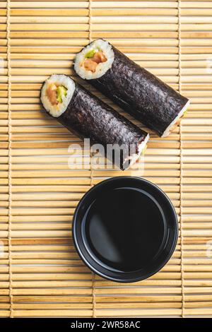 Due sushi roll e una ciotola di salsa di soia sono disposti ordinatamente su un tappetino di bambù. I sushi Rolls sono pieni di ingredienti freschi e pronti per essere accolti Foto Stock