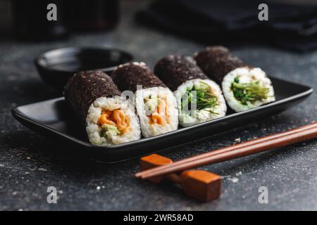 Un piatto nero con pezzi di sushi con bacchette e ciotola di salsa di soia. Il sushi mostra una varietà di colori, texture e ripieni, in vetrina Foto Stock