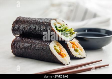 Un piatto pieno di una varietà di sushi roll con un set di bacchette di legno e una ciotola di salsa di soia, collocata su un semplice tavolo da cucina. Foto Stock