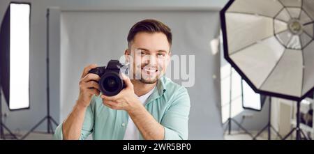 Felice fotografo sorridente che tiene una macchina fotografica in piedi in studio professionale con attrezzature leggere. Foto Stock
