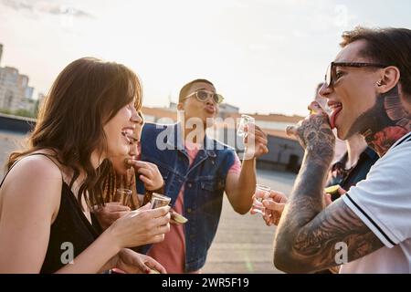 allegra gente interrazziale in abiti vivaci che si gode la tequila con sale e calce durante la festa sul tetto Foto Stock