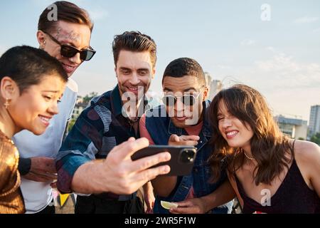 amici interrazziali gioiosi in abiti informali che guardano le foto sullo smartphone alla festa sul tetto Foto Stock