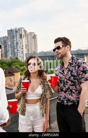 amici piacevoli e variegati con tazze rosse e bevande che parlano felici alla festa sul tetto Foto Stock