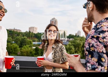 amici interrazziali gioiosi in un abbigliamento casual con occhiali da sole che bevono da tazze rosse sul tetto Foto Stock