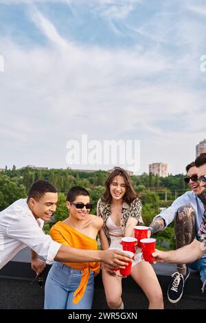 allegri amici interrazziali con gli occhiali da sole che brandono le tazze rosse con bevande mentre si trovano sul tetto Foto Stock