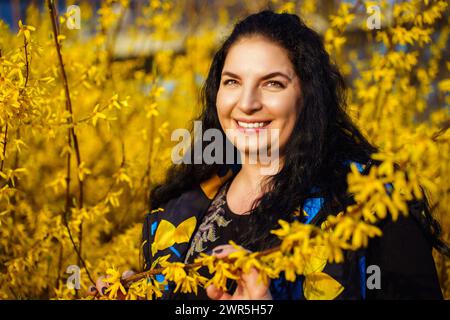 Felice giovane donna dai capelli scuri e taglia che sorride felicemente in primavera tra cespugli di forsythia gialli fioriti Foto Stock