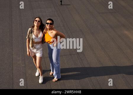 donne allegre e attraenti in vivaci abiti informali che posano insieme sul tetto e sorridono alla macchina fotografica Foto Stock