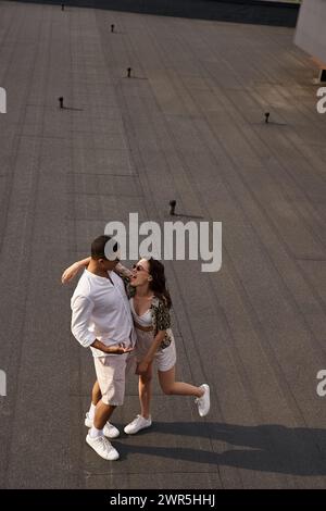 felice uomo e donna multirazziale in abiti casual con occhiali da sole in posa insieme sul tetto, coppia Foto Stock