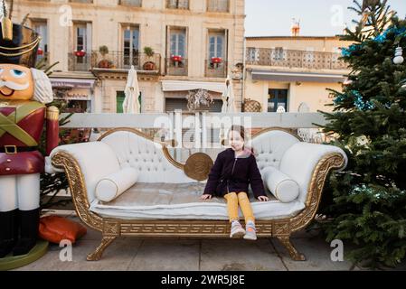 La bambina siede sul divano decorato di Natale al mercato di dicembre Foto Stock