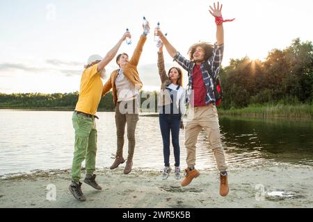 Un quartetto animato di amici che brinda vicino a un lago sereno, con il sole che tramonta dietro di loro. Amici esuberanti che brindano sulla riva del lago durante il tramonto. Foto di alta qualità Foto Stock