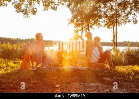 Questa immagine calda e invitante cattura un momento sereno di cameratismo e relax tra un gruppo diversificato di persone riunite in riva al lago al tramonto. Il sole, che si innalza appena sopra l'orizzonte, inonda la scena di una tonalità dorata, simboleggiando la fine di una giornata e il prezioso tempo trascorso con gli amici. I delicati raggi del sole filtrano attraverso gli alberi, creando una retroilluminazione morbida ed eterea che avvolge il gruppo in un'atmosfera accogliente. Gli individui sono vestiti in modo confortevole, suggerendo un'attività all'aperto informale o un rifugio nella natura, consentendo una pausa dal trambusto della vita quotidiana. Ogni p Foto Stock