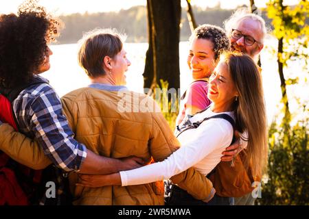 Questa immagine trasuda calore e cameratismo mentre un gruppo di amici multietnici condivide un abbraccio di gruppo sul lago durante il tramonto. I loro volti sorridenti e la vicinanza del loro abbraccio riflettono forti legami e felicità. La retroilluminazione del sole che tramonta crea un effetto alone intorno a loro, aggiungendo un senso di gioia e pace al raduno. Il variegato gruppo, compresi uomini e donne di età e background etnici diversi, illustra l'inclusività e la bellezza dell'amicizia. Gruppo abbracciato tra amici multietnici vicino al lago Suns. Foto di alta qualità Foto Stock